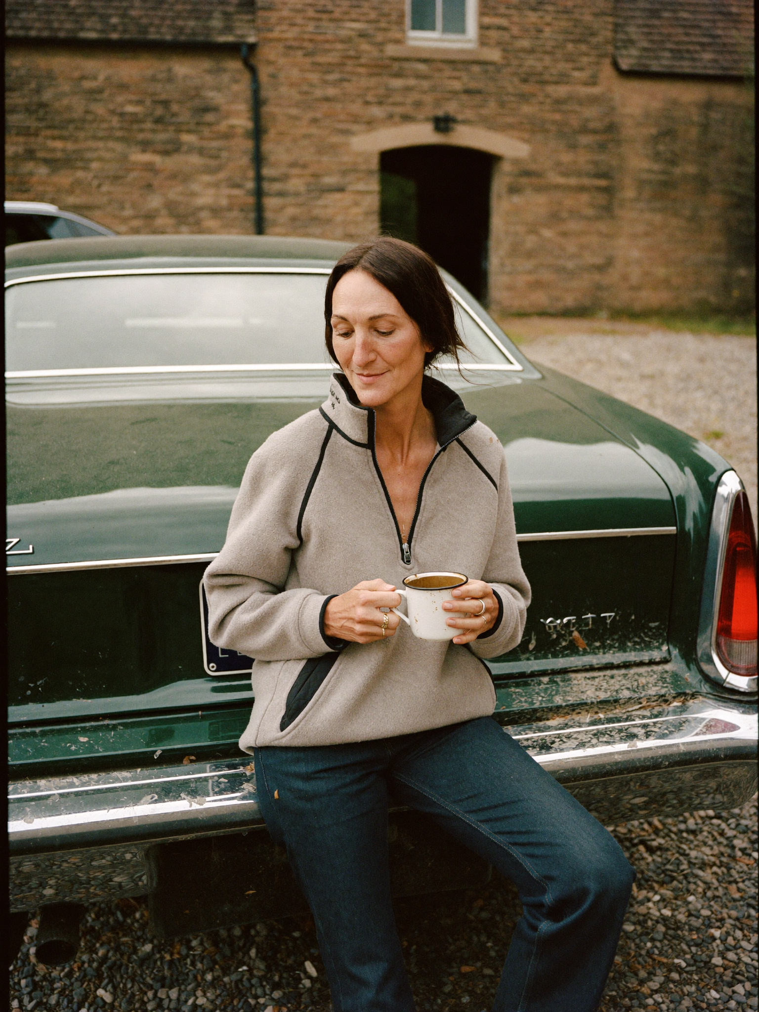 Woman in an oatmeal half-zip fleece sitting on a car tailgate in a farmyard holding a mug of tea