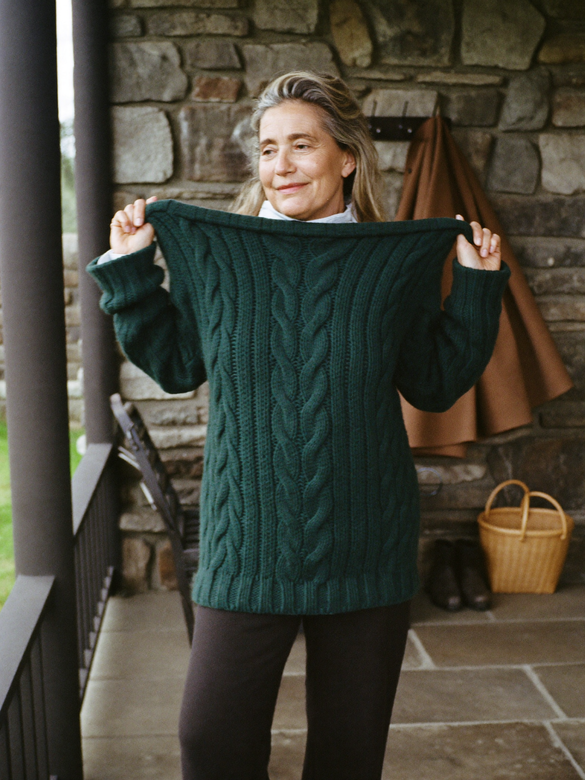 Woman pulling on an oatmeal cable knit jumper in a stone porch