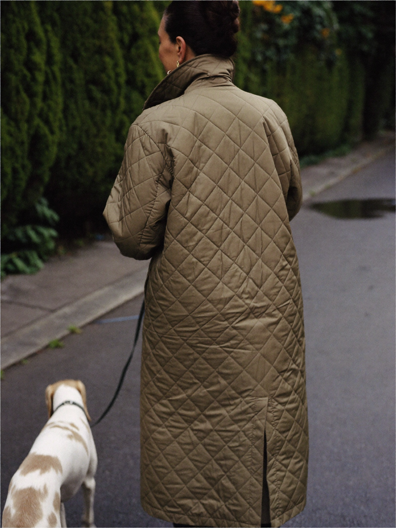 Woman walking a dog in a long olive diamond quilted coat seen from behind