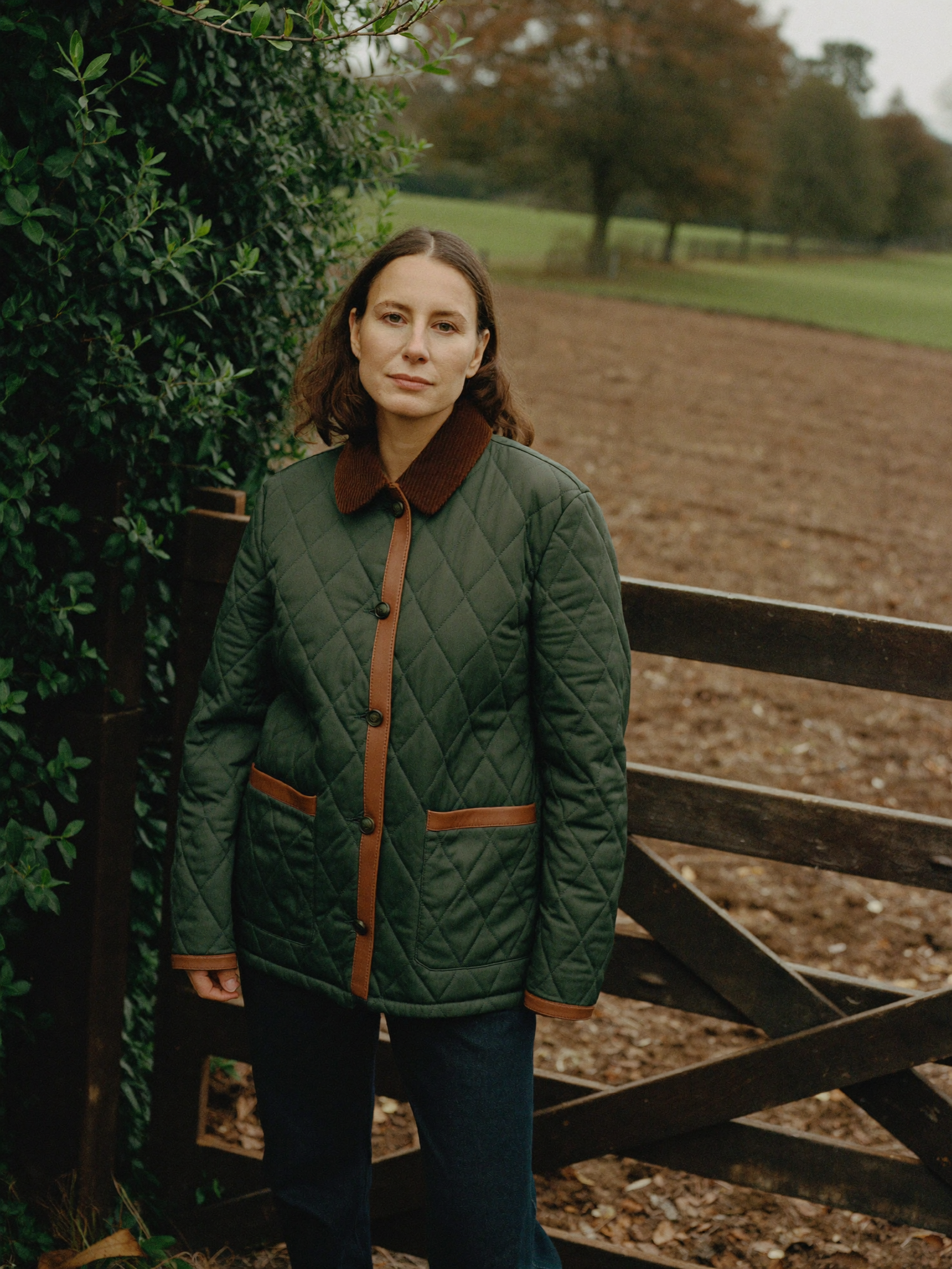 Woman in an olive diamond-quilted jacket with corduroy collar beside a five-bar farm gate