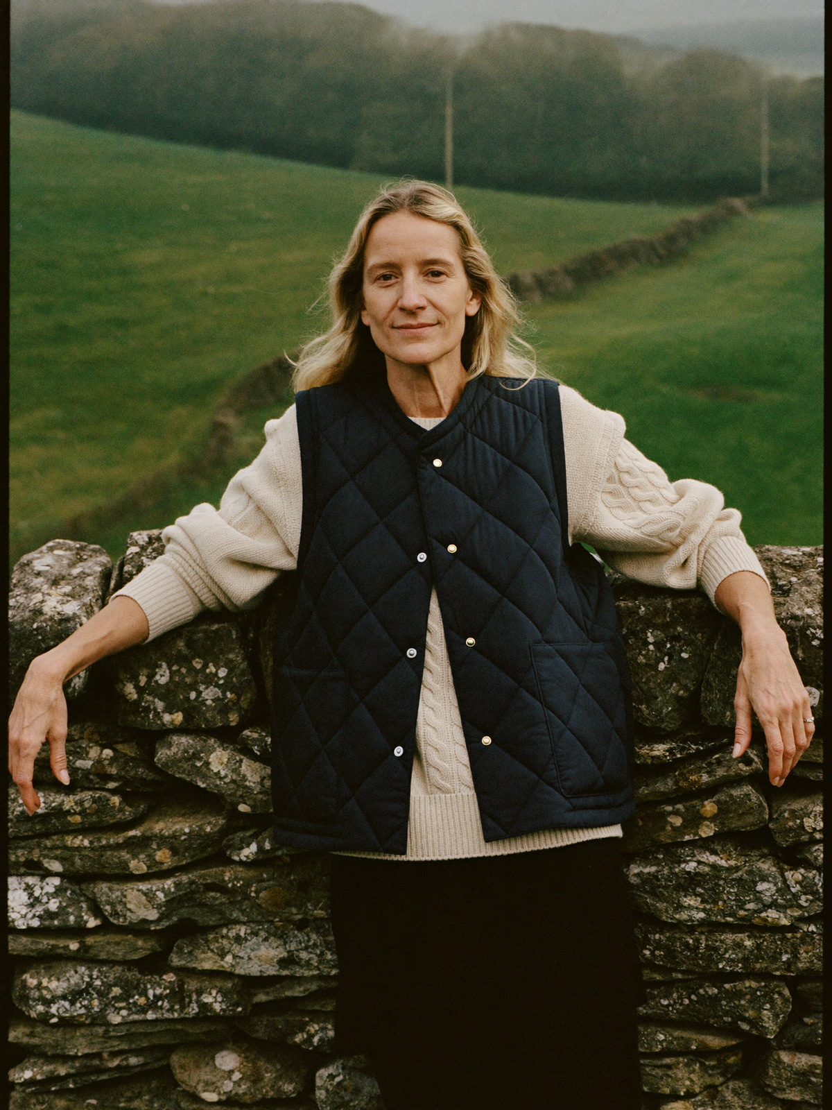 Woman in a navy quilted gilet over a cream cable knit jumper against a dry stone wall