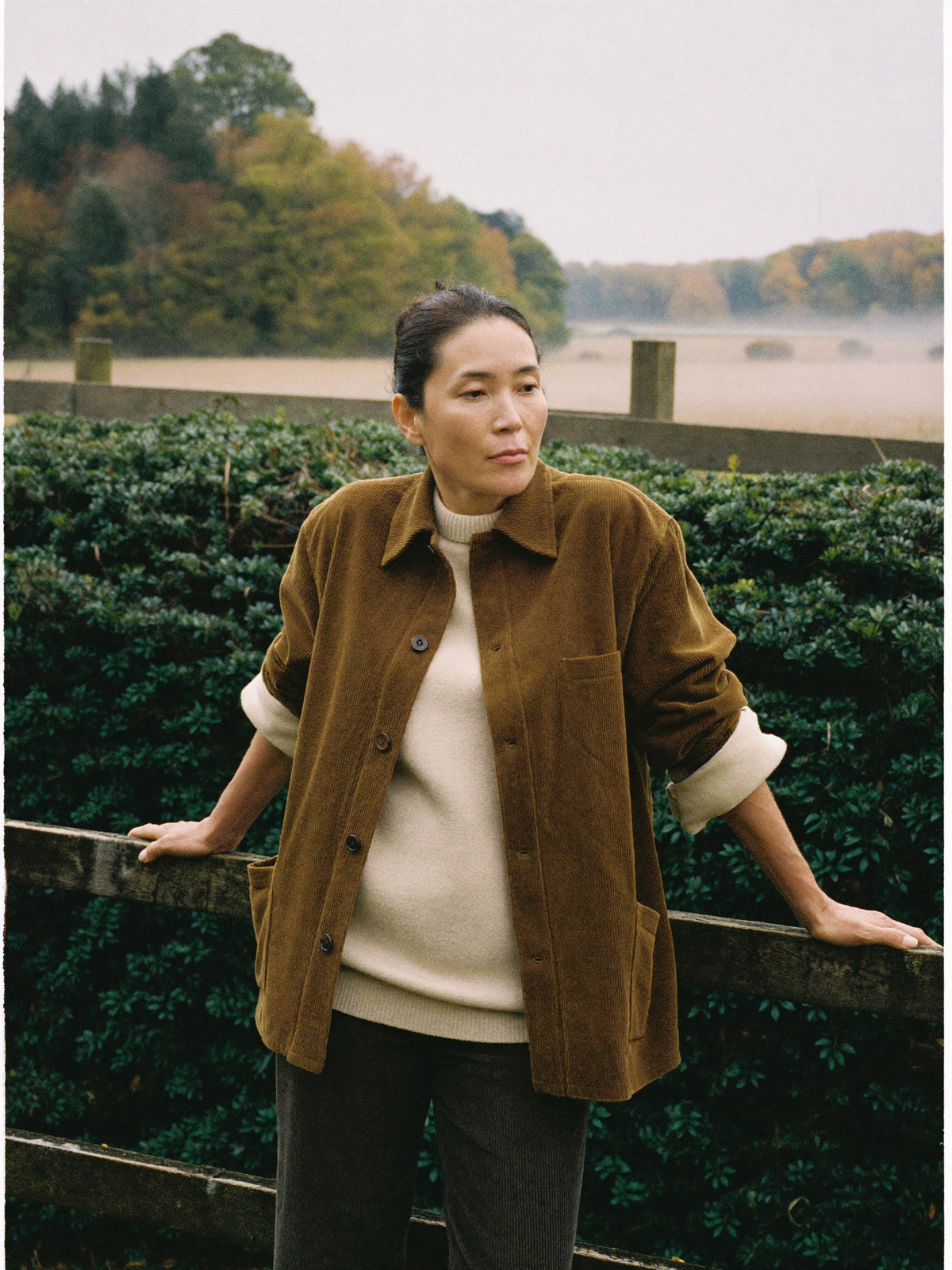Woman in a brown corduroy overshirt leaning on a wooden fence