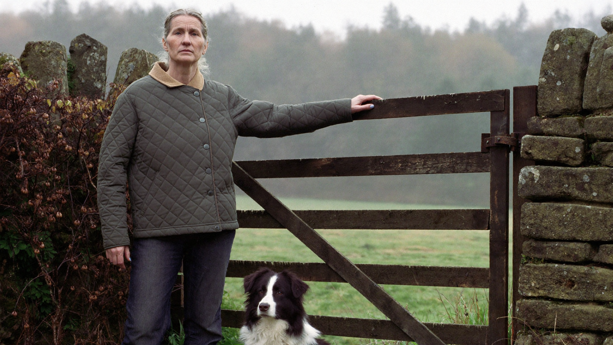 Woman in her fifties at a farm gate with a border collie, wearing an olive diamond quilted jacket with a corduroy collar